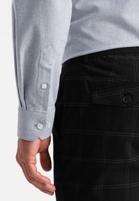 Camisa de manga larga azul claro con puños abotonados, combinada con pantalones negros con un patrón de cuadrícula y detalle en los bolsillos laterales.