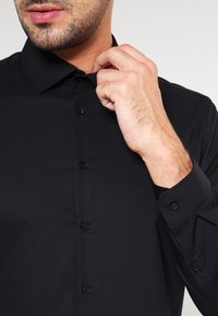 Black button-up shirt with a collar, featuring a smooth texture and long sleeves. The model adjusts the collar with one hand.