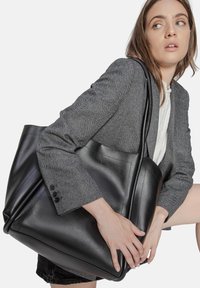 Zwarte leren schoudertas met een gladde textuur en een gestructureerd ontwerp. Kenmerkt zich door een minimalistische stijl en een contrasterende grijze herringbone blazer.