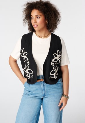 Femme aux cheveux bouclés portant un gilet noir avec une broderie florale blanche sur une chemise blanche et un jean bleu clair, regardant sur le côté.