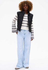 Czarna pikowana kamizelka z wysokim kołnierzem na szarym i białym swetrze w paski; szerokie jasnoniebieskie dżinsy; beżowe buty z delikatnym połyskiem.