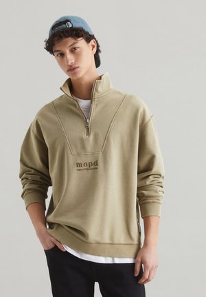 Oliwkowy sweatshirt zapinany na zamek z wysokim kołnierzem, z haftowanym logo, ściągaczami na rękawach i białą koszulą pod spodem.