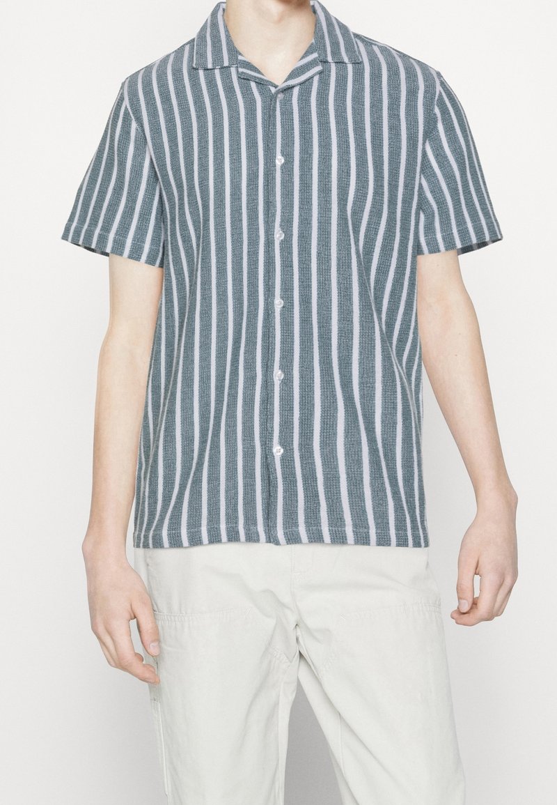 Pruhovaná košile s krátkým rukávem v barvě tyrkysové a bílé, s límcem, knoflíkovým zapínáním, strukturovanou tkaninou a vertikálními pruhy.