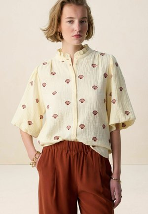 Femme portant un chemisier crème texturé avec des imprimés de coquillages rouges, devant boutonné, manches courtes bouffantes, et un pantalon taille haute couleur rouille.