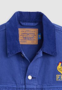 Μπλε βελούδινο σακάκι με γιακά με ετικέτα Levi Strauss & Co. και κίτρινο έμβλημα κόκορα με πατς "F.F.F." στο στήθος.
