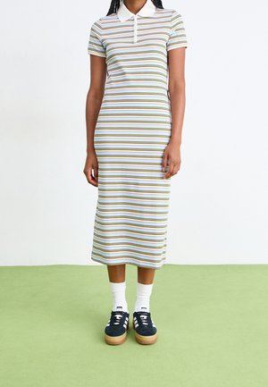 Mujer de pie sobre suelo verde, vistiendo un vestido tipo polo a rayas, de manga corta, hasta la mitad de la pantorrilla, con calcetines blancos y zapatillas negras.