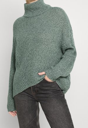 Femme portant un pull à col roulé vert en grosse maille avec une main dans la poche d'un jean gris foncé sur un fond uni.