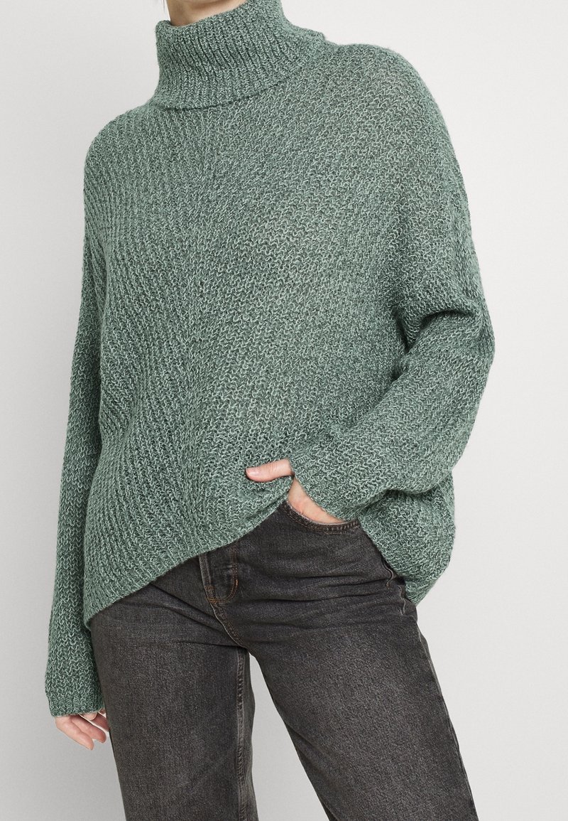 Femme portant un pull à col roulé vert en grosse maille avec une main dans la poche d'un jean gris foncé sur un fond uni.