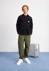 Svart manchester skjorta med knappstängning och frampocket, kombinerad med olivgröna cargobyxor och vita sneakers. Metallisk resväska i bakgrunden.