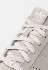 Vaaleanharmaa mokkanahkainen tennari, jossa on teksturoitu pinta, pyöreät kermaiset nauhat, rei'itetyt sivut ja logopatch kielen kohdalla.