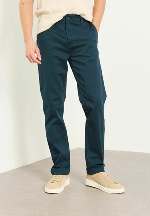 Mann trägt dunkelgrüne gerade geschnittene Hose mit beigefarbenen Sneakers und cremefarbenem Pullover, steht auf einem hellgrauen Boden vor einer weißen Wand.