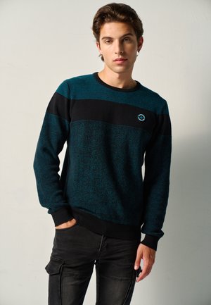 Junger Mann mit kurzen braunen Haaren, der einen türkis-schwarz gestreiften Pullover und schwarze Cargohosen trägt, steht mit einer Hand in der Tasche.