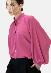 Blusa rosa brillante con botones en la parte delantera, mangas oversized y una textura suave y lisa. Estilizada con pantalones negros.