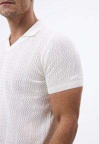 Camisa polo de punto blanca con cuello en V, que presenta un patrón texturizado y mangas cortas. Los puños y el cuello acanalados realzan el diseño.