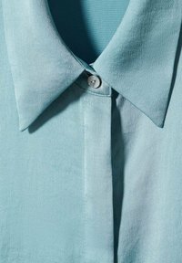 Lyseblå skjorte med krave lavet af glat stof, med knaplukning og subtilt mønster på overfladen.