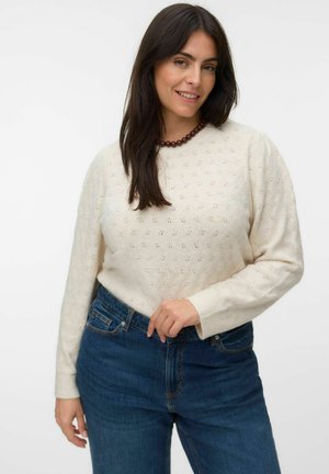Femme aux cheveux foncés portant un pull en maille blanche, un jean bleu et un collier marron en perles sur un fond blanc uni.