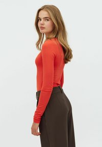 Langsleeve fitted top in felrode stof, gecombineerd met hoge taille donkere bruine broek, die een gestroomlijnd silhouet benadrukt.