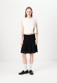 Krém színű ujjatlan felső, fekete, redőzött szoknya lyukacsos részletekkel, és fényes fekete loaferek fehér zoknival. Egyszerű, modern design.