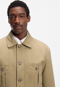 Bēša viegla virsjaka ar pogām priekšpusē, divām krūšu kabatām un izplestu apkakli. Gluda tekstūra un minimālistisks dizains.