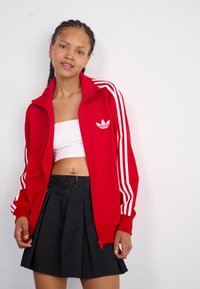 Rote Sportjacke mit weißen Streifen und Logo, kombiniert mit einem schwarzen plissierten Rock. Glatte Textur und taillierte Passform.