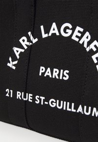 Musta kangaskassi, jossa valkoiset tekstit: "KARL LAGERFELD," "PARIS," ja "21 RUE ST-GUILLAUM." Teksti on paksua ja erottuvaa kangasta vasten.