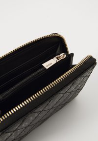 Porte-monnaie zippé noir avec un design matelassé, fermeture éclair en ton doré et quincaillerie en métal avec le logo "GUESS". L'intérieur comprend une poche zippée.