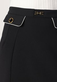 Sort nederdel med struktureret overflade, der har hvid piping, en guldknapdetalje og et geometrisk spænde ved taljen.