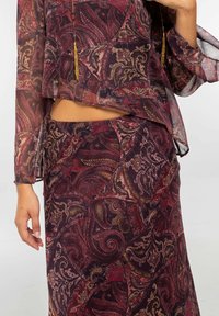 Blusa de manga larga transparente en borgoña con estampado de paisley, con cintura corta y falda de talle alto a juego, complementada con pendientes de borlas doradas.