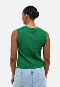 Pull en tricot vert avec un motif texturé, de design sans manches et col en côtes. Le matériau semble doux et respirant.