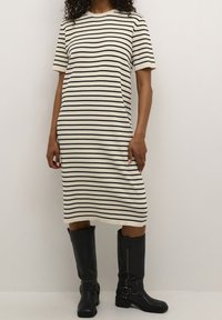 Robe en maille avec des rayures noires horizontales sur fond crème, manches courtes, et coupe décontractée. Portée avec de grandes bottes noires.