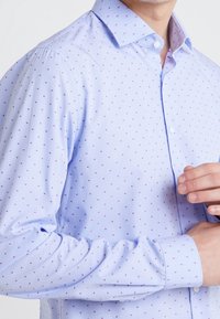Camisa de botón azul claro con un pequeño patrón de puntos rosas repetidos. La tela tiene una textura suave con un cuello y puños estructurados.