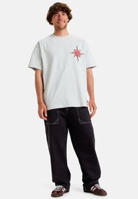 Light gray cotton T-shirt with a red sun graphic on the chest, paired with black loose-fitting pants and brown sneakers.