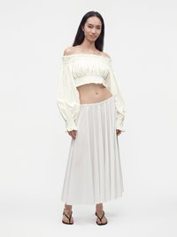 Γυναίκα που φοράει λευκό crop top με έξω τους ώμους και μακριά μανίκια και μια λευκή, αέρινη, ψηλόμεση midi φούστα με μαύρα σανδάλια.