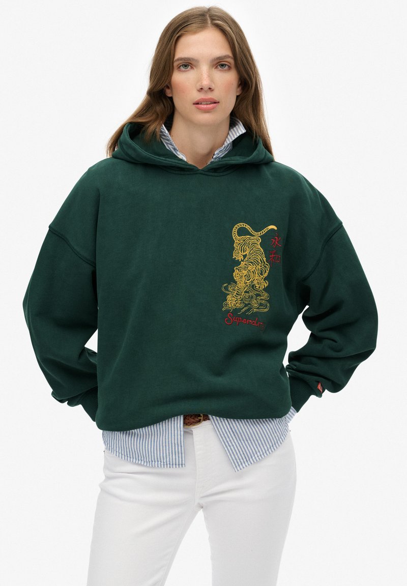 Vrouw draagt een donkergroene hoodie met een geel tijgerontwerp en rode tekst, daaroverheen een blauw gestreept shirt en witte broek.