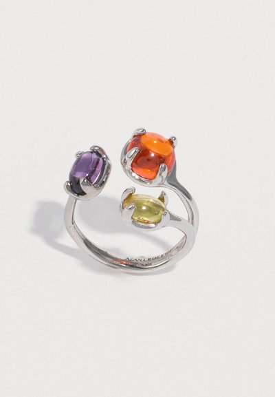 ασημένιο δαχτυλίδι με τρεις οβάλ πολύτιμους λίθους: μοβ, πορτοκαλί και κίτρινο. Η λεία μεταλλική μπάντα περιστρέφεται γύρω, αναδεικνύοντας τους πολύχρωμους λίθους.