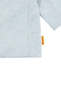 T-shirt in cotone azzurro chiaro con maniche corte e un'etichetta laterale con sfondo giallo e logo di un orso. Tessuto morbido, design casual.