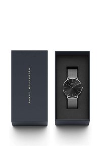 Reloj negro con correa de malla plateada en una caja azul oscura. El reloj presenta una esfera minimalista con agujas y logotipo plateados. El interior de la caja es negro.