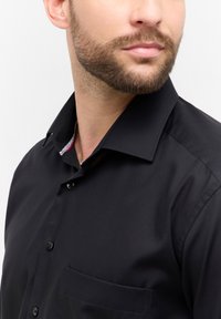 Camicia nera con bottoni e taschino, caratterizzata da un colletto strutturato e da un dettaglio interno del colletto con motivo. Tessuto liscio e vestibilità sagomata.