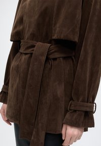 Veste en daim marron ceinturée avec manches longues et sangles aux poignets portée par une personne avec une main visible et un pantalon noir.