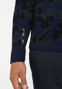 Gros plan sur une main et une manche avec un motif de camouflage bleu marine, vert et noir, et une broderie blanche "RLX" sur le poignet.