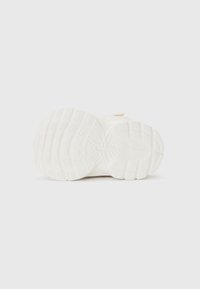 Semelle de chaussure blanche avec des motifs de grip texturés, comportant des sections circulaires et rectangulaires surélevées pour une meilleure traction. Design épuré, sans marques notable.