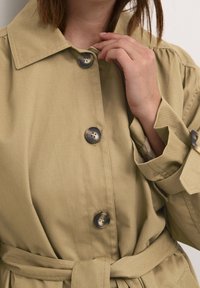 Be�żowy trencz z dużym kołnierzem, zapięciami na guziki i paskiem w talii. Charakteryzuje się gładką fakturą i widocznymi detalami szwów.