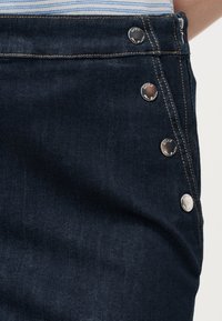 Σκούρο μπλε τζιν παντελόνι με ασημένια κουμπιά κατά μήκος της πλαϊνής ραφής και αντίθετη ραφή, φορεμένο με μια ριγέ πουκάμισο που είναι μπασιμομένη μέσα.