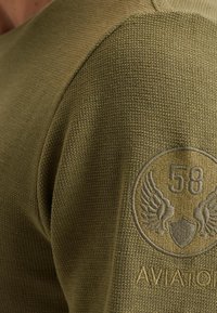 Maslinasto zelena pletena dukserica s teksturiranom tkaninom; ima kružni izvezeni pečat s krilima i brojem 58 na rukavu.