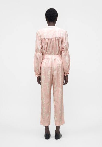 Άτομο που στέκεται γυρισμένο με την πλάτη, φορώντας ταιριαστό ροζ μακρυμάνικο πάνω μέρος και παντελόνι με φωτεινό λουλουδάτο σχέδιο και μαύρα παπούτσια.