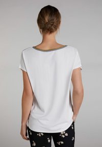 Top blanco de manga corta con un escote redondeado que presenta acentos en negro y dorado. La espalda es lisa, con un corte holgado.