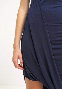 Kobieta ma rękę opartą na boku, ubrana w ciemnoniebieską drapowaną spódnicę lub sukienkę na neutralnym tle.