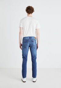Jeans in denim blu con vestibilità dritta, tasche posteriori con dettagli di cucitura e una patch in cuoio marrone sulla vita. Abbinati a una camicia bianca.