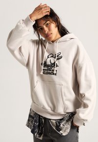 Light beige hoodie with front pocket; features a large graphic design and text. Layered with a dark printed shirt underneath.