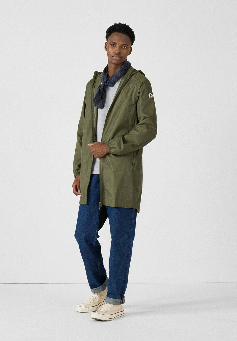 Parka imperméable vert olive avec une capuche, fermeture éclair, poignets élastiques et un écusson logo sur la manche, associé à un pantalon en denim décontracté et des baskets.